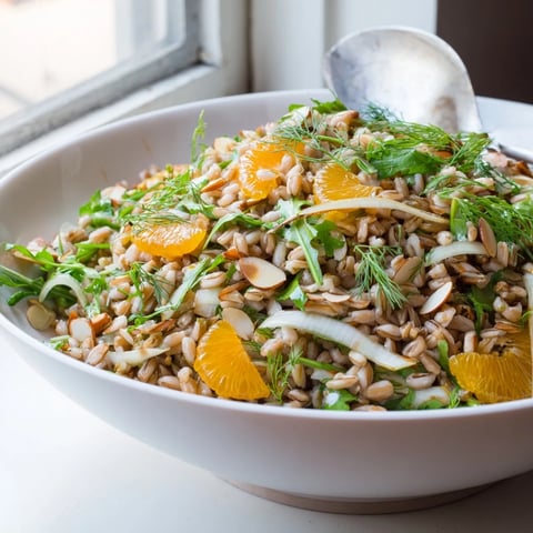 A vibrant bowl of Farro Salad With Fennel, Oranges, and Almonds topped with fresh parsley and fennel fronds on a wooden table.